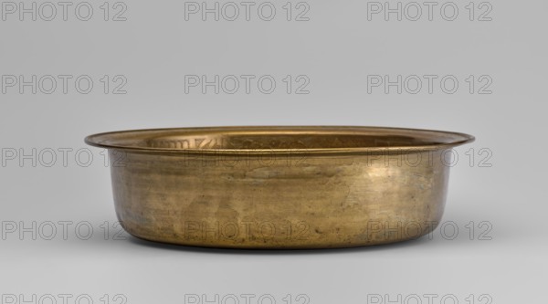 Bowl, fruit, images of birds, bowls, plant ornament, Chinese art, Chinese culture, peaches fruits, lingzhi, pumpkins, pomegranates fruits, happiness symbol, deer, artemisia, cranes, pine trees, Chinese symbols. Round bowl with collar collar decorated with a plant tendril. 19th/20th century, China. Vessel; height 8.7 cm, diameter 33 cm., A shallow, round metal basin with a wide flared rim sits centered against a neutral background; the basin appears made of a warm golden-bronze metal showing areas of darker patina and subtle irregular marks and scratches on its exterior surface, with a slightly reflective inner surface visible along the rim. The background and surface beneath the basin are light gray, and the overall colors visible are gold, bronze, brown, gray, and hints of darker tarnish.