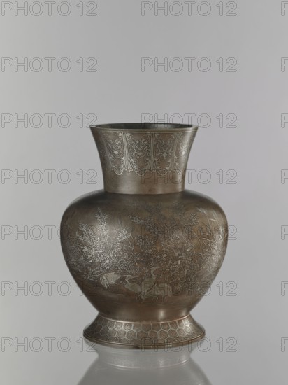 Vase, pewter alloy ovoid bulbous vase on a wide base with a wide flared neck. Engraved decoration inlaid with a thin layer of silver: cranes among reeds by the water; a symmetrically repeated floral motif on the neck; kikko pattern; grasses; birds. Meiji period 1868–1912, 19th/20th century, Japan. Vessel. Height 30.1 cm; diameter 15.6 cm., A metal vase with a flared neck and rounded body standing on a slightly flared base, rendered in bronze tones with areas of darker and lighter brown and subtle metallic gray highlights; the neck features a repeating decorative band with ornate leaf-like motifs in a lighter metallic tone, the body is engraved with a natural scene showing a group of birds wading among detailed grasses and flowering plants in a paler metallic shade, and the base bears a geometric honeycomb-like pattern in similar bronze and gray hues.