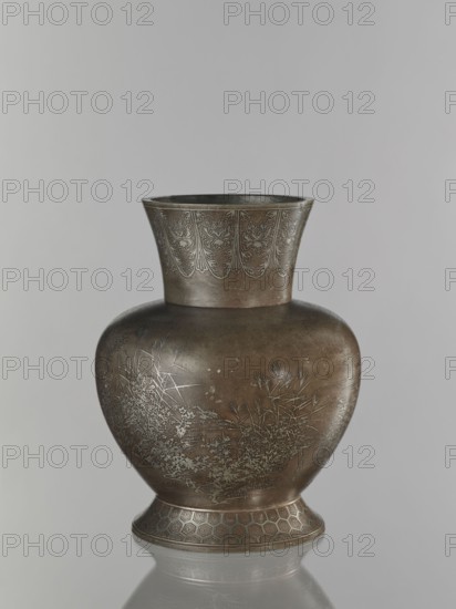 Vase, pewter alloy, ovoid bulbous vase on a wide base with a wide flared neck. Engraved decoration inlaid with a thin layer of silver: cranes among reeds by the water; a symmetrically repeated floral motif on the neck; kikko pattern; grasses; birds. Japanese, Meiji period 1868–1912, 19th/20th century. Vessel. Height 30.1 cm, diameter 15.6 cm., A bulbous metal vase with a flared rim sits against a neutral gray background, showing a patinated surface in shades of brown, bronze, and muted green; the upper rim area has a band of engraved decorative motifs in a lighter metallic tone, while the body displays etched scenes of grasses and foliage in a silvery metallic color; the base flares outward and is decorated with a repeating geometric pattern in bronze and darker brown, and the vase casts a soft reflection on the surface below.