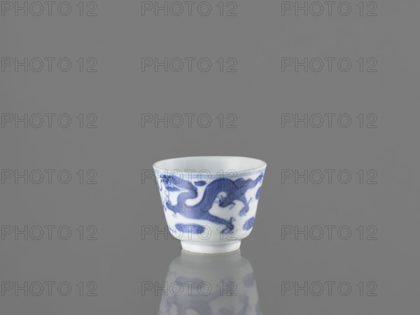Bowl, gift, fenghuang iconography, Chinese art, dragons iconography, Chinese culture, birds, phoenixes iconography, blue and white porcelain, 19th century China, vessel, height 4.5 cm, diameter 5.7 cm, A small white porcelain cup with a blue painted dragon motif around its exterior sits on a reflective gray surface against a solid gray background, showing the cup's rounded shape, flared rim, and the cup's reflection beneath it; visible colors include white, various shades of blue, and gray.