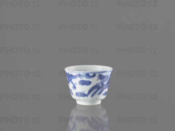 Bowl, ; blue-and-white porcelain vessel from China, 19th century; height 4.5 cm, diameter 5.7 cm. Decoration: dragons, fenghuang/phoenixes, birds; Chinese art., A small white porcelain cup with blue painted designs sits centered on a reflective gray surface against a plain gray background, the cup showing a flared rim and a short foot, and its blue decoration forming stylized organic shapes and figures that are mirrored faintly in the reflection; visible colors include white, blue, and various shades of gray.