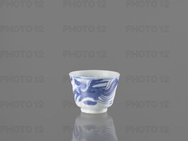Bowl, ; fenghuang phoenix and dragons iconography; Chinese art; birds; blue-and-white porcelain; 19th century China; vessel; height 4.5 cm, diameter 5.7 cm, A small white ceramic cup with blue painted flowing designs sits centered on a reflective smooth surface, showing a faint mirrored reflection beneath it; the background is a uniform medium gray, and the cup's blue decoration features wave-like and organic shapes wrapping around its exterior.