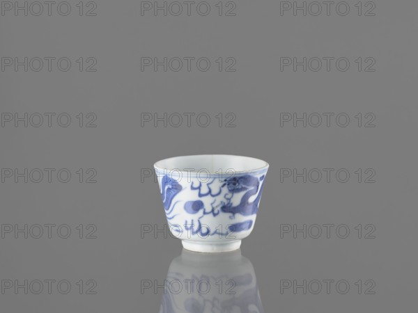 Bowl, fenghuang iconography, Chinese art, dragons iconography, Chinese culture, birds, phoenixes iconography, blue-and-white porcelain, 19th century China, vessel, height 4.5 cm, diameter 5.7 cm, A small porcelain cup sits centered on a smooth reflective surface against a gray background, showing a white base color with blue painted decorative shapes and brushstrokes around its exterior; the cup has a slightly flared rim and a short foot, and its reflection is visible below, with colors including white, blue, and various shades of gray.