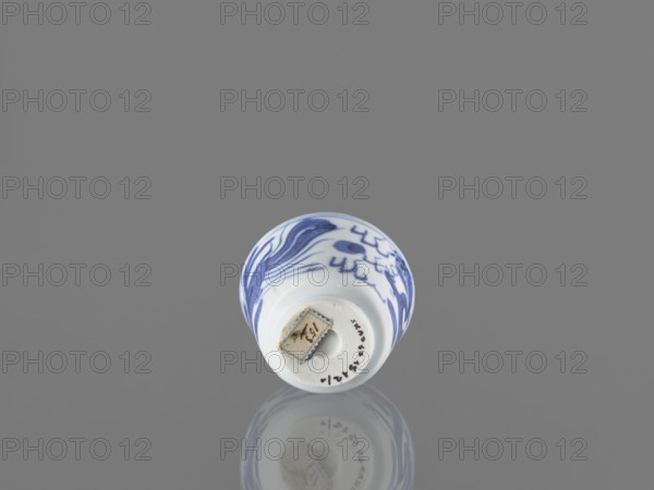 Bowl, fenghuang iconography, Chinese art, dragons iconography, Chinese, birds, phoenixes iconography, blue and white porcelain, 19th century, China, vessel, height 4.5 cm, diameter 5.7 cm, A small ceramic cup viewed from above with a rounded rim and a narrow base, decorated with blue brushstroke patterns on a white surface, resting on a reflective grey surface that creates a soft mirrored image below; visible colors include blue, white, grey, and a small patch of beige on the cup's base.