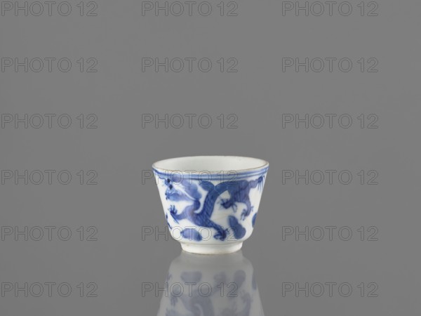 Bowl, Chinese, 19th century. Blue-and-white porcelain vessel height 4.6 cm; diameter 5.9 cm with dragons, fenghuang/phoenixes and birds iconography, decorated with pearls., A small white ceramic cup with blue painted decorative motifs sits centered on a reflective surface against a plain gray background, the cup showing a rounded rim that tapers slightly toward a narrow base and its blue painted design appearing as flowing, organic shapes around the exterior, with the cup and its blue-and-white reflection visible on the surface.