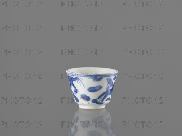 Bowl, Chinese, 19th century. Blue-and-white porcelain vessel decorated with dragons, phoenixes fenghuang, and birds; includes pearl motifs. Height 4.6 cm, diameter 5.9 cm., A small white ceramic cup with blue painted abstract and organic brushstroke patterns sits centered on a reflective gray surface against a plain gray background, showing a soft reflection below; the cup has a slightly flared rim and a narrow foot, with visible blue decoration around its exterior and a white interior.