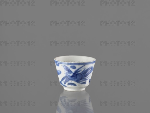 Bowl, Chinese, 19th century. Blue-and-white porcelain vessel decorated with dragons, phoenixes fenghuang, and birds; includes pearl motifs. Height 4.6 cm; diameter 5.9 cm., A small white porcelain cup with blue painted decoration sits on a reflective gray surface against a gray background, showing a stylized blue bird motif and abstract blue brushstrokes around its exterior and a narrow blue rim at the top, with a subtle reflection of the cup visible on the surface.