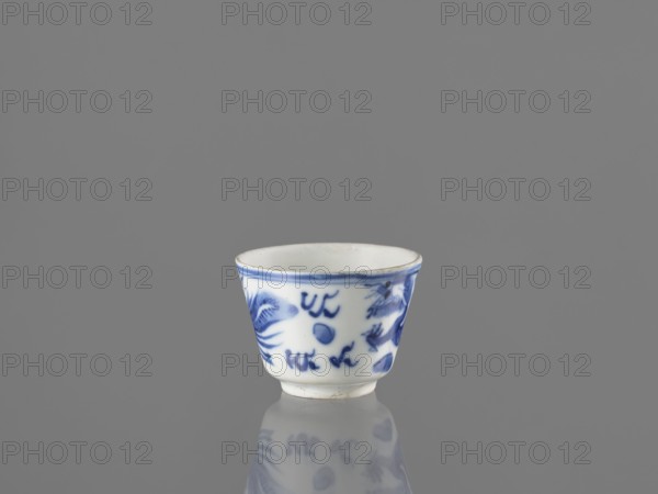 Bowl, Chinese, 19th century. Blue-and-white porcelain vessel decorated with dragons, phoenixes fenghuang, and birds; pearls motif. Height 4.6 cm, diameter 5.9 cm., A small ceramic cup with a white base and blue painted designs sits on a reflective surface, showing a faint mirrored reflection below; the cup has a slightly flared rim with a thin blue band, blue brushstroke motifs around the exterior that resemble abstract shapes and floral elements, and a smooth white interior; the background and surface are shades of gray.