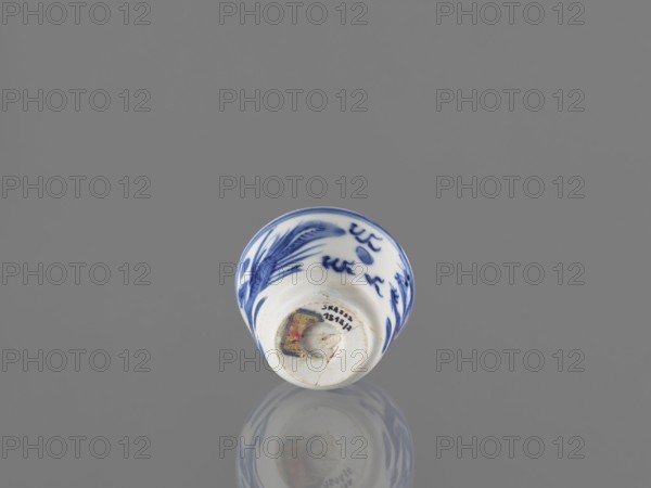 Bowl; Chinese; 19th century. Blue-and-white porcelain vessel decorated with dragons, phoenixes fenghuang, and birds; pearls motif. Height 4.6 cm, diameter 5.9 cm., A small porcelain cup sits on a reflective gray surface, showing blue painted brushwork on a white background with organic, flowing motifs and a blue rim; the cup is viewed from a low angle revealing its rounded interior and base, and its underside and a fragment of colored material are visible, with the cup's reflection mirrored faintly on the surface.