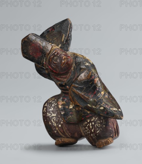 Netsuke in the shape of a sanbasô dancer; sculptor, Japanese, Edo period 18th century; material: carved netsuke figurine; subject: dance/ritual dancer wearing a mask; dimensions: height 6.3 cm, width 4.0 cm., A carved wooden figure with worn paint showing a group of dark colors including black, brown, red, gold, white, and hints of green, depicted in a crouching posture with rounded legs and a tilted upper body; the surface shows chipped and flaked areas revealing layers of pigment and exposed wood, with carved facial features on a rounded head and a pointed element projecting from the back or shoulder area; the object rests on a smooth neutral gray background and casts a soft shadow beneath it.