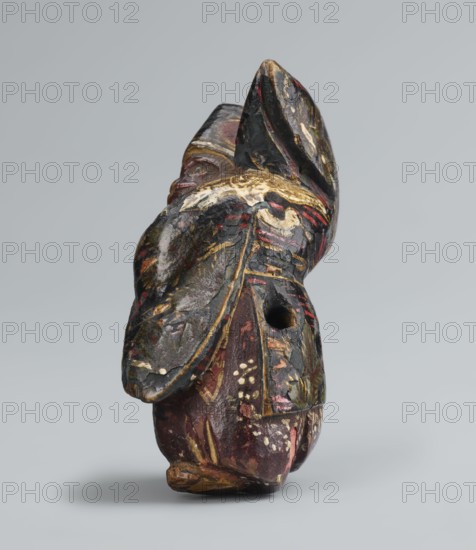 Netsuke in the shape of a sanbasô dancer, Japanese, Edo period 18th century, netsuke figurine depicting dance/ritual dancer with mask, height 6.3 cm, width 4.0 cm., A small carved object with a worn surface showing a mix of dark colors including black, deep red, brown, and gold, with areas of pale cream where the surface is chipped; the object has a rounded base and an irregular upper portion with layered, folded shapes and carved ridges, and a visible round hole on one side; the painted surface shows decorative brushlike marks and speckled pale dots, and the piece casts a soft shadow on a plain light gray background.