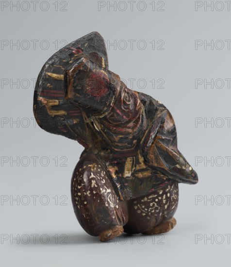 Netsuke in the shape of a sanbasô dancer, Japanese, Edo period 18th century, figurine netsuke depicting dance/ritual dancer with mask; height 6.3 cm, width 4.0 cm., A carved wooden figure with worn paint, showing a seated or crouching posture and a large headpiece or hat, rendered in dark colors including black, deep brown, red, gold, and touches of beige and green; the surface has visible brushstrokes and areas of chipped or cracked paint revealing layers beneath; the base consists of rounded forms with speckled beige markings, and the figure's carved facial features and folded limbs show textured, angular carving; the background is a smooth pale gray.