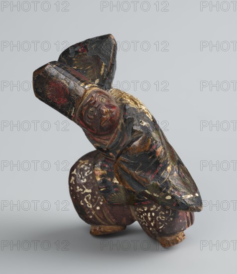 Netsuke in the shape of a sanbasô dancer;, Japanese, Edo period 18th century; material: carved netsuke figurine; subject: ritual dance, dancer wearing a mask; dimensions: height 6.3 cm, width 4.0 cm., A small carved figure with a rounded face and pronounced features wearing a tall hat is shown in a crouching pose; the surface shows worn paint in black, deep red, muted gold, brown, and touches of green, with areas of flaked and chipped finish revealing lighter wood beneath, and patterned white speckles on the rounded lower portion, all set against a plain light gray background.