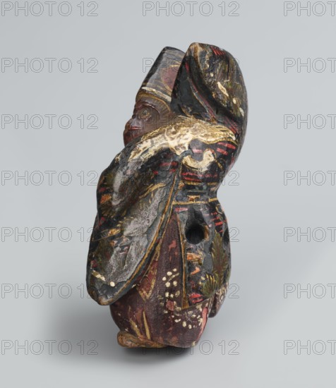 Netsuke in the shape of a sanbasô dancer, Japanese, Edo period 18th century, netsuke figurine depicting a ritual dancer with mask, height 6.3 cm, width 4.0 cm., A carved figurine viewed from an angle showing its side and back, with visible wear and layered paint. The surface shows dark tones including black and deep brown, along with red and maroon areas, patches of gold and yellowish highlights, touches of white, and small green accents. The figure has a rounded head and a prominent curved element that wraps around the back, textured and chipped in places, and there is a visible circular hole on one side. The base is irregular and the paint appears cracked and rubbed, revealing underlying material in a few spots.