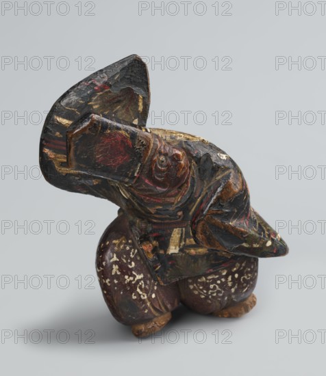 Netsuke in the shape of a sanbasô dancer, Japanese, Edo period 18th century, netsuke figurine depicting dance/ritual dancer wearing a mask; height 6.3 cm, width 4.0 cm., A small carved wooden figure with a rounded body and an oversized head is shown against a pale gray background; the object displays worn layers of dark brown and black paint with patches of red, gold, and muted green, and areas where the paint has chipped to reveal lighter wood tones; the carving shows curved, rounded shapes and incised details around what appears as facial features and a beaklike projection, with a textured, aged surface and scattered flecks of off-white on the lower portion.