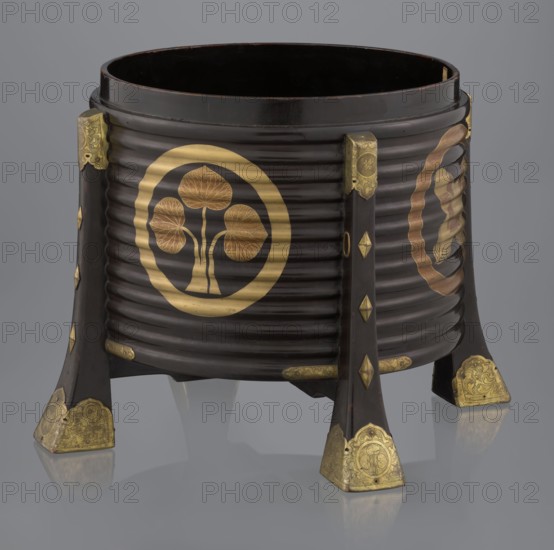Container hokai for shells for the game kai awase, Japanese, Meiji 1868–1912 and Edo 1603–1868 periods. Wood covered with black lacquer, decorated with gold maki-e lacquer, gilded copper fittings. Decorated with feathers and aoi hollyhock plant ornament. 19th century Edo–Meiji, Japan, Edo. Container; height 37.5 cm, diameter 40.0 cm., A dark brown cylindrical wooden container with a ribbed exterior standing on a few flared legs, each leg tipped with decorative metallic corner pieces in a golden color; the body of the container features several circular golden motifs with stylized organic shapes inside, and there are additional small golden decorative accents along the legs and rim; the interior appears dark brown to black, and the background is a smooth grey.