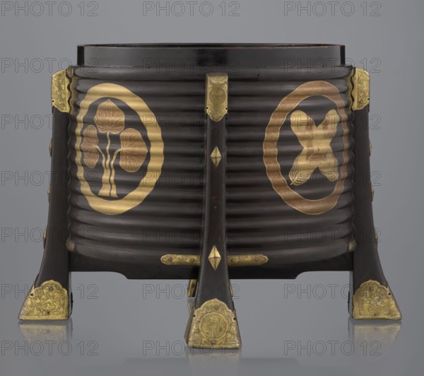 Container hokai for shells for the game kai awase, Japanese, Meiji 1868–1912 and Edo 1603–1868 periods. Wood covered with black lacquer, decorated with gold maki-e lacquer, gilded copper fittings. Decoration: feathers, aoi hollyhock plant ornament. Function: container for shells used in the game kai awase. 19th century Edo–Meiji period, Japan; Edo. Height 37.5 cm; diameter 40.0 cm., A dark brown, ribbed cylindrical container elevated on a tripod of three dark brown legs with ornate gold-colored metal fittings at the feet and connecting points; the main body shows two large circular gold-colored emblems on its front face, one containing stylized leaf-like shapes and the other containing a different symmetrical motif, both set against the dark brown ribbed surface; the legs have small gold-colored decorative accents along their length and gold-colored corner protectors with engraved patterns; the top rim is dark brown and the background behind the object is a neutral gray.