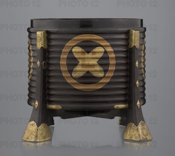 Container hokai for shells for the game kai awase;, Japanese; Meiji 1868–1912 and Edo 1603–1868 periods; wood covered with black lacquer, decoration with gold maki-e lacquer, gilded copper fittings; decorated with feathers and aoi hollyhock plant ornament; boxes and shells for parlor/game use; 19th century Edo–Meiji; place of origin: Edo, Japan; container; height 37.5 cm, diameter 40.0 cm., A dark brown cylindrical wooden container with horizontal ridges stands on four flared legs; the container features several gold-colored metal fittings and ornamentation at the top corners, along the legs, and on the foot ends, and a large circular gold-colored decorative motif is visible on the front; the legs have diamond-shaped gold-colored accents and the foot caps have engraved patterns; the background is a smooth gray gradient and the surface beneath reflects the object.
