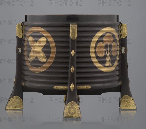 Container hokai for shells for the game kai awase, Japanese, Meiji 1868–1912 and late Edo 1603–1868 periods, wood covered with black lacquer, decoration with gold maki-e lacquer, gilded copper fittings; decorated with feathers and aoi hollyhock plant ornament; boxes, shells, parlor game; 19th century; Edo, Japan; container; height 37.5 cm, diameter 40.0 cm, A dark brown, ribbed cylindrical object set on four angled legs with decorative metal fittings; the surface shows gold circular motifs on the front sides featuring stylized organic shapes and a crossed ribbon-like form, and several gold ornamental plates and diamond-shaped accents on the legs and central support. The visible colors are dark brown, gold, and a neutral gray background.