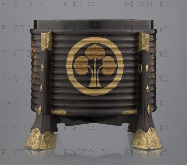 Container hokai for shells for the game kai awase;, Japanese; Meiji 1868–1912 and Edo 1603–1868 periods; materials: wood covered with black lacquer, decoration with gold maki-e lacquer, gilded copper fittings; decoration: feathers, aoi hollyhock plant ornament; function: boxes for shells used in parlor game kai awase; 19th century Edo–Meiji; place of origin: Edo, Japan; dimensions: height 37.5 cm, diameter 40.0 cm., The image shows a dark brown cylindrical wooden object with horizontal ridges and a circular gold emblem featuring a stylized three-leaf plant on its front. The piece rests on four dark brown legs with decorative gold metal fittings at the top and bottom of each leg, including ornate gold caps at the feet. The wood has a polished, glossy appearance and the gold details contrast with the dark brown surface. The background is a smooth neutral gray and the object casts a faint reflection on the glossy surface beneath it.