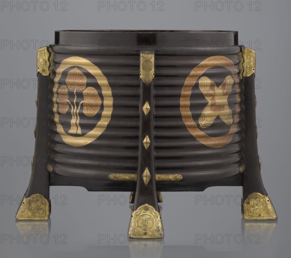 Container hokai for shells for the game kai awase, Japanese, Meiji 1868–1912 and late Edo 1603–1868 periods, wood covered with black lacquer, decoration with gold maki-e lacquer, gilded copper fittings. Decorated with feathers and aoi hollyhock plant ornament. Function: container for shells used in the parlor game kai awase. 19th century Edo–Meiji. Place of origin: Edo, Japan. Height 37.5 cm; diameter 40.0 cm., A dark brown ribbed wooden container with a round top supported by four angled legs, decorated with gold metal fittings on the leg ends and on the top corners; the front shows a pair of circular gold emblems, one with a stylized tree-like motif and the other with an abstract symmetrical motif, both rendered in gold tones with some reddish-gold shading; the legs and central vertical support feature small gold diamond-shaped accents and engraved gold plates with floral patterns; the background is a smooth gray.