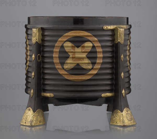 Container hokai for shells for the game kai awase;, Japanese; Meiji 1868–1912 and Edo 1603–1868 periods; wood covered with black lacquer, decorated with gold maki-e lacquer, gilded copper fittings; decorated with feathers and aoi hollyhock plant ornament; boxes, shells, parlor game object; 19th century Edo–Meiji; Japan, Edo; container; height 37.5 cm, diameter 40.0 cm, A dark brown, ribbed cylindrical container resting on a stand with flared legs, decorated with several gold-colored metal fittings and ornaments; the front shows a large circular gold-colored emblem with an X-shaped motif inside; the legs and corners have ornate gold-colored corner plates and diamond-shaped gold-colored accents, and there are gold-colored horizontal embellishments near the base; the overall colors visible are dark brown, gold, and a neutral gray background.