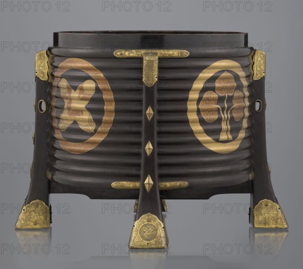 Container hokai for shells for the game kai awase;, Japanese; Meiji 1868–1912 and Edo 1603–1868 periods; wood covered with black lacquer, decorated with gold maki-e lacquer, gilded copper fittings; decorated with feathers and aoi hollyhock plant ornament; used for boxes, shells, and parlor game kai awase; 19th century Edo–Meiji; place of origin: Edo, Japan; container; height 37.5 cm, diameter 40.0 cm, A dark brown, ribbed wooden container with a curved front stands on a set of short legs with gilded metal fittings; the front shows a central vertical wooden brace with small gilded diamond-shaped ornaments and elaborately engraved gold-colored metal plates at the top and bottom, and a few circular gold-colored medallion-like designs are set into the ribbed sides. On the left side of the front, inside a gold-colored oval, there is a stylized gold motif with curved, ribbon-like elements, and on the right side, inside a matching gold-colored oval, there is a gold motif resembling clustered ...