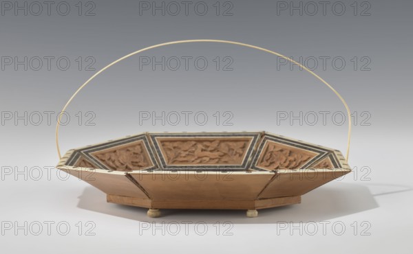 Basket. Octagonal shallow wooden basket with an ivory rim and handle. Inside the basket is a relief scene: in the central field a palace surrounded by rich vegetation, at the foot of which a wild, spotted animal leopard? tiger? dog? strolls. A trail of stars is carved around this scene. The side panels alternate with images of the same animal and flowers; spaces between panels are filled with colorful mosaic tiles. Handle and rim are ivory. Served both utilitarian and decorative purposes. 19th century, India; height 14.0 cm, width 24.0 cm; floral and animal ornamentation., A shallow, octagonal wooden basket with a thin, curved pale yellow handle rising above it, showing carved decorative panels inside the sloping sides; the basket displays natural wood tones, pale beige, light brown, dark brown, and black accents, and rests on small round feet casting soft shadows on a light gray surface beneath a gradient gray background.