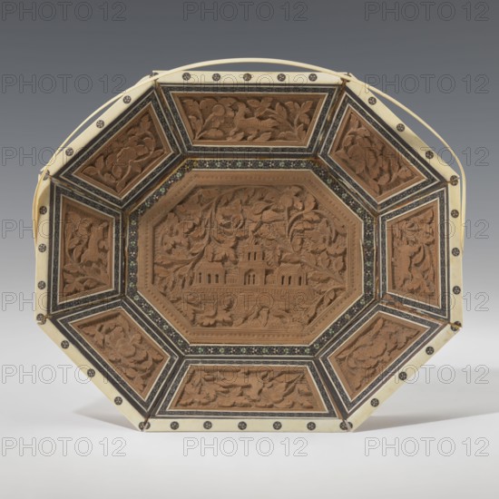 Basket. Octagonal, shallow wooden basket with an ivory rim and handle. Inside the basket is a relief scene. In the central field is a palace surrounded by rich vegetation, at the foot of which a wild, spotted animal leopard? tiger? dog? strolls. A trail of stars is carved around this scene. The basket's side panels alternate with images of the same animal and flowers. The spaces between the panels are filled with colorful mosaic tiles. The basket's handle and rim are made of ivory. The basket served both utilitarian and decorative purposes. 19th century, India. Container; height 14.0 cm, width 24.0 cm. Indian art; South Asian; floral ornament; animals; flowers; plant ornaments; mosaics; leopards., An octagonal carved wooden tray with a central raised panel depicting a building surrounded by floral and foliate motifs, framed by a decorative band with small inlaid pieces and a border of alternating dark and light geometric bands; each side panel contains carved floral scenes and animal f...