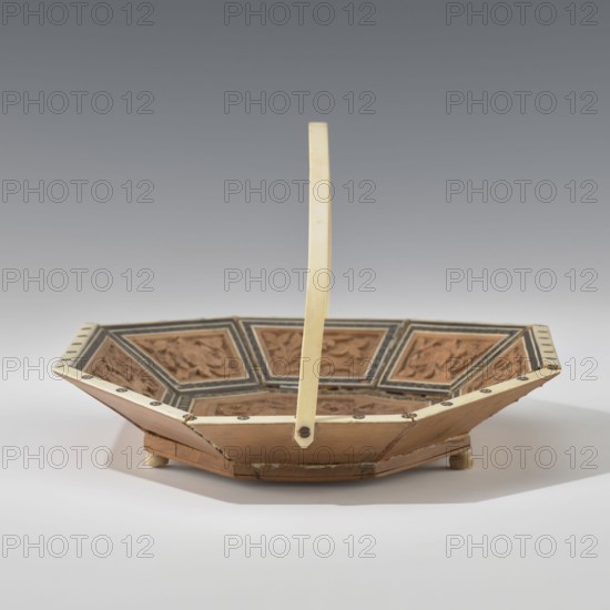 Basket, Octagonal, shallow wooden basket with an ivory rim and handle. Inside the basket is a relief scene: in the central field a palace surrounded by rich vegetation, at the foot of which a wild, spotted animal leopard? tiger? dog? strolls. A trail of stars is carved around this scene. The basket's side panels alternate with images of the same animal and flowers. The spaces between the panels are filled with colorful mosaic tiles. The basket's handle and rim are made of ivory. The basket served both utilitarian and decorative purposes. 19th century, India; container. Height 14.0 cm, width 24.0 cm., An octagonal, shallow basket-like object with a tall, thin vertical handle rising from its center, showing a combination of beige, tan, brown, black, and cream colors; the interior features several recessed panels with carved or molded floral or foliate relief in brown tones framed by darker borders, while the rim and handle are a lighter cream or ivory color accented by small dark circula...