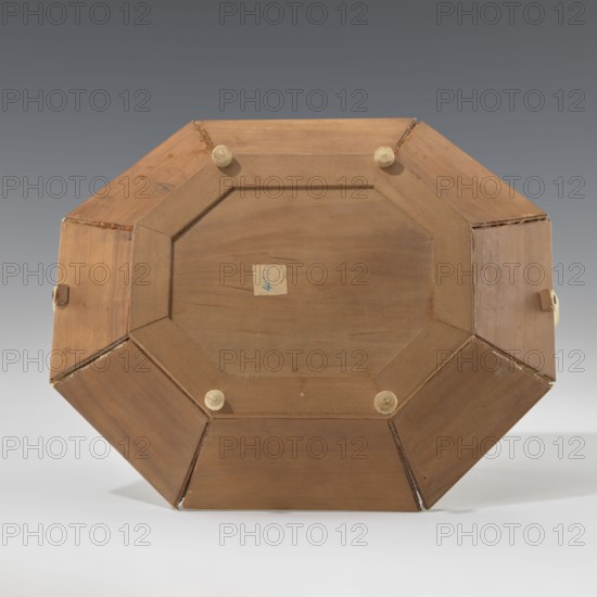 Basket, gift, Indian art, Indian culture, floral ornament, South Asian culture, architecture, animals, containers, flowers, plant ornaments, mosaics, leopards. Octagonal, shallow wooden basket with an ivory rim and handle. Inside the basket is a relief scene: in the central field is a palace surrounded by rich vegetation, at the foot of which a wild, spotted animal leopard? tiger? dog? strolls. A trail of stars is carved around this scene. The basket's side panels alternate with images of the same animal and flowers. The spaces between the panels are filled with colorful mosaic tiles. The basket's handle and rim are made of ivory. The basket served both utilitarian and decorative purposes. 19th century, India; container; height 14.0 cm, width 24.0 cm., A wooden octagonal object viewed from one flat side, showing a central recessed panel surrounded by angled wooden segments with visible grain and joins, a group of small round beige feet or pegs attached near the inner edge, a few small ...
