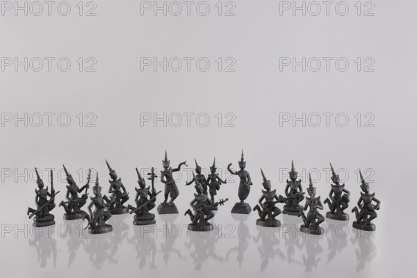 Set of fourteen figurines of musicians, Southeast Asia, art of Southeast Asia, music, dance, musical instruments, 1st half of the 20th century, Cambodia, figurine sculpture, height 10–13 cm, A row of small dark metallic figurines arranged on a reflective light surface, each figure wearing pointed headdresses and posed in various stances with a few holding instruments or objects, their reflections visible below them; the background is a pale off-white to light gray expanse.