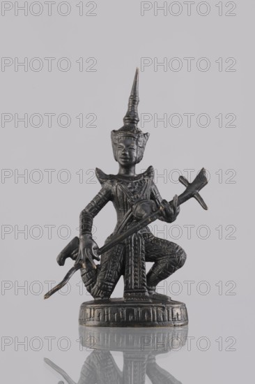 Musician figurine, Southeast Asian art, Southeast Asia culture, musicians, headgear, 1st half of the 20th century, Cambodia place of origin, sculpture, figurine, height 11.1 cm, A small dark metallic figurine of a kneeling figure wearing an ornate pointed headdress and detailed patterned clothing, holding a long curved object across the body; the figure stands on a round decorated base and shows textured surface details and slight worn highlights in a few areas, with colors including dark gray and metallic goldish accents against a pale gray background and a reflective surface below that mirrors the base and parts of the figure.