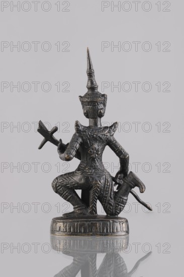 Musician figurine, Southeast Asian art, Southeast Asia culture, musicians, headgear, 1st half of the 20th century, Cambodia, sculpture, figurine, height 11.1 cm, A small dark metallic figurine is shown from the back, kneeling on a round base with ornamental patterns; the figure wears an ornate pointed headdress and detailed armor with carved patterns, holding a cross-shaped object in one hand and a curved object in the other; the surface shows a few worn highlights of a lighter metallic color against the dominant dark tone, and the figurine casts a faint reflection on the pale gray background.