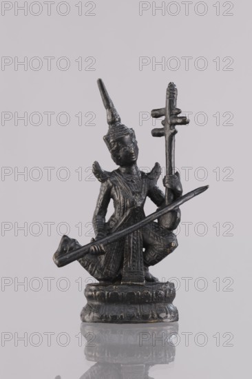 Musician figurine, Southeast Asia culture, Southeast Asian art, musicians, 1st half of the 20th century, Cambodia place of origin, figurine, sculpture, height 10.9 cm, A small dark metal figurine stands on a round pedestal, showing a kneeling figure wearing a tall conical headdress and detailed clothing with textured patterns; the figure holds a long straight object diagonally across its body and supports a vertical staff with horizontal crosspieces in one hand, and small ornamental wing-like shapes appear at the shoulders. The surface of the figurine is primarily dark gray to black with subtle lighter wear spots, and it is set against a pale gray background with a faint reflective surface beneath that shows a soft mirrored silhouette.