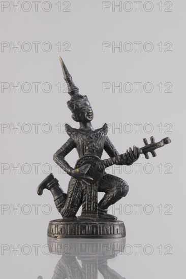 Musician figurine, Southeast Asian art, musicians, Southeast Asia culture, 1st half of the 20th century contemporary product, Cambodia, sculpture, figurine, height 10.9 cm, A small dark metallic figurine with a few areas of worn golden highlights shows a kneeling figure wearing an ornate pointed headdress and patterned clothing, holding a stringed instrument with a rounded body and a long neck; the figure is positioned on a circular decorated base and casts a faint reflection on a smooth light gray surface, with the background a plain pale gray.