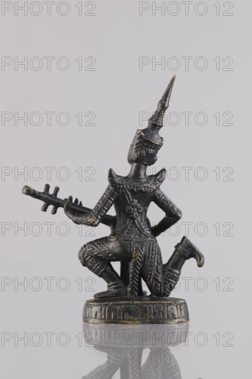 Musician figurine, Southeast Asian art; musicians; Southeast Asia culture; unknown provenance; 1st half of the 20th century contemporary product; Cambodia place of origin; sculpture; figurine; height 10.9 cm, A small dark metallic figurine with worn gold highlights depicts a kneeling figure wearing ornate patterned clothing and a tall pointed headdress, holding a long, toothed instrument across its body; the figure rests on a round base with decorative edges, and the scene is reflected on a glossy surface against a pale gray background with tones of white and light gray.