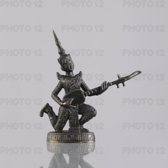 Musician figurine, Southeast Asian art, musicians, Southeast Asia culture, 1st half of the 20th century contemporary product, Cambodia place of origin, sculpture, figurine, height 11.1 cm, A small dark metal figurine with hints of gold patina depicting a kneeling figure wearing a tall pointed headdress and ornate clothing, holding a long curved instrument or object across its lap and outstretched in one hand; the figure stands on a round decorated base that reflects faintly on the smooth light gray surface beneath it.