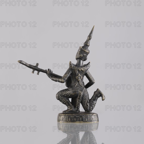 Musician figurine, Southeast Asian art, musicians, Southeast Asia culture, provenance unknown, 1st half of the 20th century contemporary product, Cambodia place of origin, sculpture, figurine, height 11.1 cm, A small dark metallic figurine with worn gold highlights shows a kneeling figure wearing an ornate tall pointed headdress and detailed patterned clothing, holding a long thin staff-like object horizontally with one arm extended; the figure is mounted on a round decorated base and sits against a plain light gray background with a soft reflection of the base visible on the surface below.