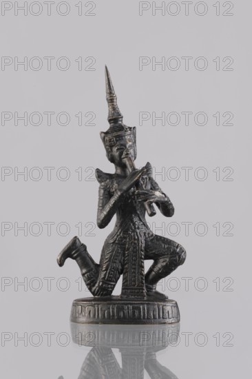 Musician figurine, Southeast Asian art, musicians, Southeast Asia culture, 1st half of the 20th century contemporary product, Cambodia, sculpture, figurine, height 11.2 cm, A small dark metallic statuette of a kneeling figure wearing an ornate tall pointed headdress, with detailed textured clothing and armor, holding a flute-like instrument to its mouth with both hands; the figure is positioned on a round decorated base and there is a soft reflection of the base and lower figure on the glossy surface below. The visible colors are dark gunmetal, black, muted bronze highlights, and pale gray in the background.