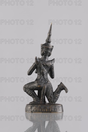 Musician figurine, Southeast Asian art; musicians; Southeast Asia culture; unknown provenance; 1st half of the 20th century contemporary product; Cambodia place of origin; sculpture, figurine; height 11.2 cm, A small dark metallic figurine with a pointed tall headdress sits on a round base, shown from the back; the figure wears detailed patterned clothing with raised decorative elements and has a knee bent as if kneeling, with textured surface areas showing worn highlights in a few places. The visible colors are dark gray to black with small areas of muted gold or bronze highlights and a neutral light gray background.