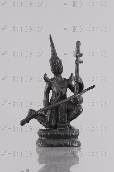 Musician figurine, Southeast Asian art, musicians, Southeast Asia culture, 1st half of the 20th century contemporary product, Cambodia place of origin, sculpture, figurine, height 11.1 cm, A small dark metallic statue of a seated figure wearing a pointed headdress, holding a long thin rod across the body and a taller vertical object in one hand, with decorative ridges and textured clothing, standing on a rounded ornamental base, against a pale gray background; visible colors include dark gray, black, and subtle metallic highlights.