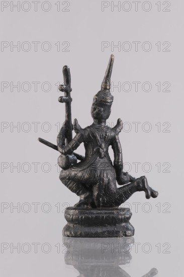 Musician figurine, Southeast Asian art; subject: musicians; culture: Southeast Asia; provenance, 1st half of the 20th century contemporary product; place of origin: Cambodia; medium: sculpture, figurine; height: 11.1 cm, A small dark metallic statue of a kneeling figure viewed from the back, with a pointed headdress and ornate textured clothing, sitting on a rounded pedestal; the figure holds a vertical staff-like object in one hand and another object that extends outward in the other hand; the surface shows subtle highlights and worn areas, and the overall colors visible are dark gray, black, and small hints of gold, set against a pale gray background with a faint reflection of the pedestal on the glossy surface beneath.