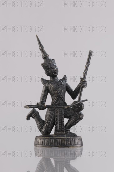 Musician figurine, Southeast Asia culture, art of Southeast Asia, 1st half of the 20th century contemporary product, Cambodia, sculpture, figurine, height 10.9 cm, A dark metallic statuette of a kneeling figure wearing an ornate pointed headdress and detailed patterned clothing is shown against a pale gray background, the figure holding a long slender rod vertically in one hand and a curved implement horizontally in the other, with textured armor-like sleeves, decorative shoulder pieces, and a flared skirt-like lower garment, standing on a round carved base that sits on a reflective surface; visible colors include dark gray to black metal with small highlights of gold and the pale gray of the background and reflection.