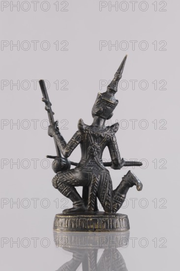Musician figurine, Southeast Asia culture, art of Southeast Asia, 1st half of the 20th century contemporary product, Cambodia, sculpture, figurine, height 10.9 cm, A small dark metallic statue of a kneeling figure with ornate patterned clothing and a tall pointed headdress, holding a long thin staff in one hand and a rounded object near the other hand; the statue sits on a decorated circular base and shows subtle worn highlights in a gold tone against the primary dark metal color, with a pale neutral background and a faint reflection of the statue visible below.