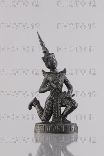 Musician figurine, Southeast Asian art, Southeast Asia culture, musicians, 1st half of the 20th century contemporary product, Cambodia place of origin, sculpture, figurine, height 11 cm, A dark metallic figurine of a kneeling human figure wearing an ornate pointed headdress and intricately patterned clothing is shown against a pale gray background, the figure holding small objects near its chest and lap, standing on a round decorated base with a subtle reflection visible on the glossy surface below; visible colors include dark gray, black, and hints of metallic gold against the light gray backdrop.