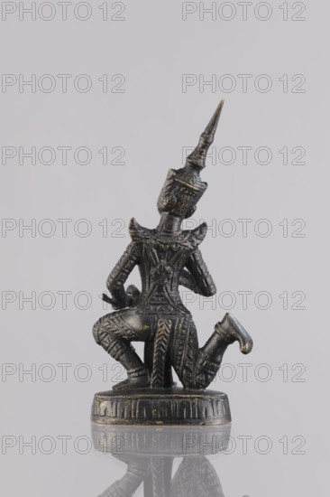 Musician figurine, Southeast Asian art, Southeast Asia culture, musicians, 1st half of the 20th century contemporary product, Cambodia place of origin, sculpture, figurine, height 11 cm, A small dark metallic figurine with subtle gold highlights showing a kneeling figure with ornate patterned clothing and armor, a tall pointed headdress, detailed arm and leg decorations, and a circular decorative base, set against a pale gray background with a faint reflection of the figurine on the glossy surface; colors present include dark gray or black, gold highlights, and pale gray.