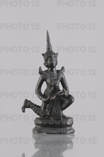 Musician figurine, Southeast Asia culture, Southeast Asian art, musicians, 1st half of the 20th century contemporary product, Cambodia, sculpture, figurine, height 11 cm, A small dark metallic figurine of a kneeling humanlike figure wearing a tall pointed headdress and decorative shoulder pieces, with a few areas showing lighter worn highlights, positioned on a rounded base that is reflected on a glossy surface, against a plain light gray background.
