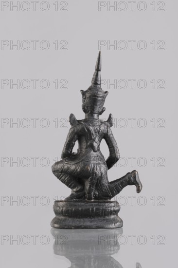 Musician figurine, Southeast Asia culture, Southeast Asian art, musicians, 1st half of the 20th century contemporary product, Cambodia, sculpture, figurine, height 11 cm, A small dark metallic figurine shown from the back, seated on a rounded base with a reflective surface beneath it, featuring a tall pointed headdress, stylized shoulders, and draped clothing; the visible colors are dark gray to black with subtle lighter highlights and a pale gray background.