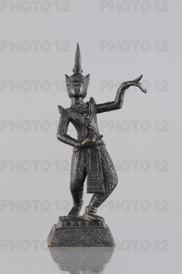 Dancer figurine, Southeast Asian art; subject: musicians; culture: Southeast Asia, Cambodia; provenance, 1st half of the 20th century contemporary product; medium: figurine, sculpture; height: 12.7 cm, A dark metallic figurine of a person in a dynamic pose stands on a textured base against a light gray background; the figure wears an ornate pointed headdress and detailed patterned clothing, with one arm raised and curved and the other bent at the waist, and the surface shows areas of darker black and subtle brownish-gold highlights.